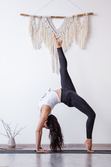 Young woman asian yoga lover doing standing in Bridge exercise, One legged Wheel pose on floor in cozy white room. Working out, wearing sportswear, black pants, bra, indoor full length, home interior