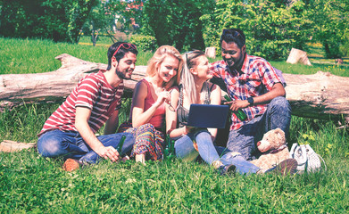 Wall Mural - Multiracial group of cheerful friends having fun with pc at city park - Happy students sitting on green grass using laptop in positive social moment at spring - Summer concept of joy youth and leisure