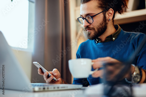 Young handsome bearded hipster sitting at his home office, holding cup of coffee and using smart phone Tableau sur Toile