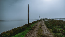 View Of A Dirt Road With A Lonely Silhouette Of A Man Walking Into The Distance Surrounded By Grassy Parts And Water On Both Sides And Wooden Poles On The Left Side, On A Misty Dull Overcast Morning