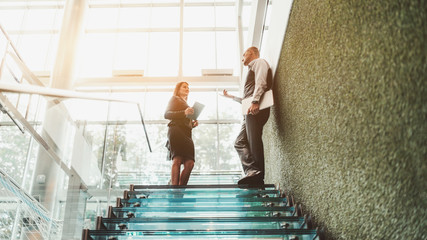 Two business people on a glass office ladder: a tall bossy caucasian man entrepreneur is giving instructions to his female subordinate, a beautiful businesswoman with a long hair and digital tablet