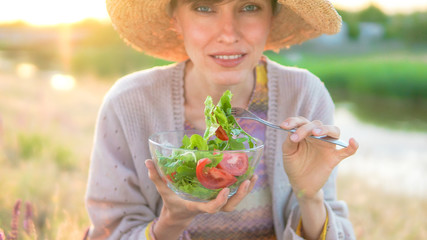 Wall Mural - Beautiful caucasian woman eating salad over green natural background with sunset or sunrise sky or light