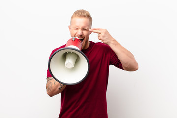 Wall Mural - young blonde man looking unhappy and stressed, suicide gesture making gun sign with hand, pointing to head against white wall with a megaphone