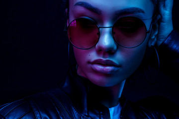 Wall Mural - portrait of an African American young woman in sunglasses isolated on a black background in neon light