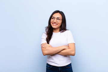 young latin pretty woman feeling happy, proud and hopeful, wondering or thinking, looking up to copy space with crossed arms against flat wall