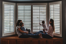 Same Sex Parents Lounging With Kids On Window Seat With Tall Shutters