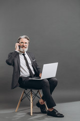 Business and fashionable bearded mature man in a gray suit taking by phone and using laptop on the grey background.