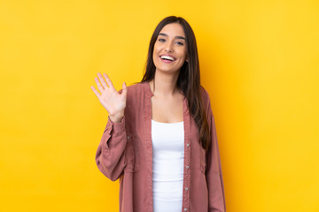 Wall Mural - Young brunette woman over isolated yellow background saluting with hand with happy expression