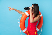 Lifeguard Woman Over Isolated Blue Background With Lifeguard Equipment And With Binoculars While Pointing Far