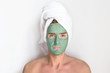 Young handsome man, metrosexual guy with cosmetic clay blue mask on his face and a towel on head looking at camera, smiling on white background. Beauty, spa, skin care concept. Men taking care of skin
