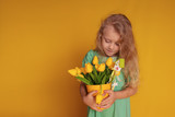 Fototapeta Tulipany - Cute cheerful girl in a green dress on a yellow background holds tulips in her hands and laughs merrily.