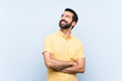 © luismolinero - Young man with beard over isolated blue background looking up while smiling
