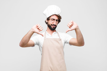 Wall Mural - young crazy chef looking sad, disappointed or angry, showing thumbs down in disagreement, feeling frustrated against white wall