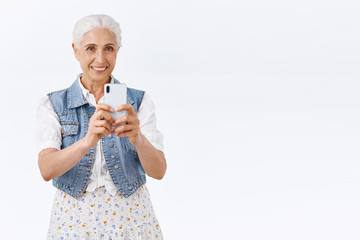 Charming modern stylish senior woman, grandmother spend time amusement park with grandkids, taking photo of kid, smiling as photographing, holding smartphone, white background