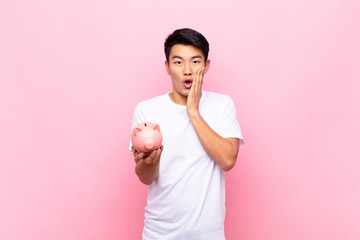 young japanese man feeling shocked and scared, looking terrified with open mouth and hands on cheeks holding a piggy bank