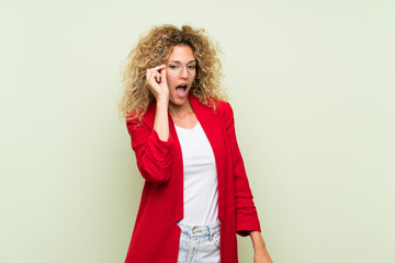 Young blonde woman with curly hair over isolated green background with glasses and surprised