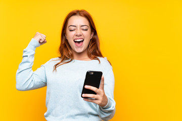 Wall Mural - teenager redhead girl over isolated yellow background with phone in victory position