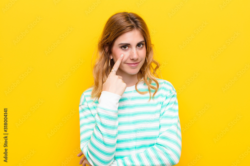 young pretty blonde woman keeping an eye on you, not trusting, watching and staying alert and vigilant against yellow wall - obrazy, fototapety, plakaty 