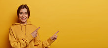 Check This Out, Better See It. Beautiful Dark Haired Millennial Girl Wears Casual Hoodie, Points Both Index Fingers On Blank Space, Has Friendly Smile, Discusses Promo, Isolated On Yellow Background.