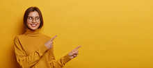 Studio Shot Of Pleasant Looking Young Woman With Dark Combed Hair, Wears Neck Sweater, Points Away On Blank Space, Demonstrates Advert, Isolated Over Yellow Background, Describes Amazing Place