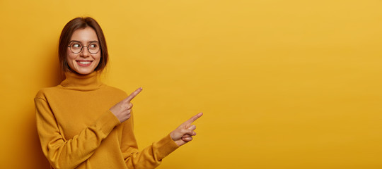 Wall Mural - Studio shot of pleasant looking young woman with dark combed hair, wears neck sweater, points away on blank space, demonstrates advert, isolated over yellow background, describes amazing place