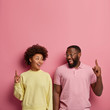 © wayhome.studio  - Vertical shot of happy dark skinned couple point fore fingers above on blank space for your advertising content, look with smile at each other, recommend going upstairs, isolated over pink background