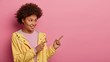 Follow this direction. Pleased smiling woman has Afro hair, points at upper right corner, suggests using copy space wisely, dressed in casual yellow anorak, advertises something over pink background