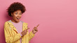 Joyful pretty young woman has curly hairstyle, points away on copy space, dressed in yellow anorak, stands against pink background, giggles positively, advertises empty space. Positive vibes.