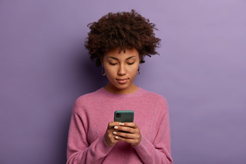 Wall Mural - Serious looking Afro American woman reads message on modern mobile phone, surfes social media, has concentrated look in display, wears casual jumper, isolated over purple wall, updates information