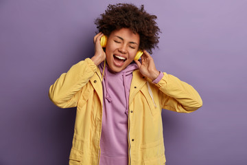 Hipster girl tilts head, sings song loudly, enjoys loud good quality sound in headphones, closes eyes, doesnt notice anybody, wears vivid yellow jacket, isolated on purple background. Feeling rhythm