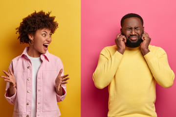 Photo of irritated woman screams with negative emotions, gestures angrily and shouts at husband who plugs ears and ignores wifes yelling, sort out realtionship, isolated on yellow and pink wall