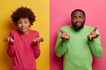 Wall Mural - Indifferent ethnic teenagers shrug shoulders and stand hesitant, make decision, choose between something, have doubts, wear bright pink and green jumpers, stand indoor. Colorful vivid background