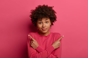 Wall Mural - Confused beautiful young African American woman crosses arms over body, points left and right, different directions, chooses best option, has various special offers, wears bright pink jumper.