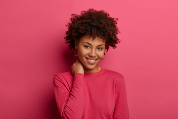 Wall Mural - Good looking woman with curly hair, keeps hand on neck, smiles gently, wears crimson casual jumper, looks gladfully at camera, isolated over pink studio wall, has optimistic view. Happiness and joy