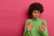 Awkward unimpressed woman with Afro hairstyle, pulls palms towards camera, refuses something, rejects proposal, wears green neck sweater, stands over crimson wall with empty space. No, thank you