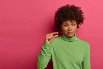 Wall Mural - Not much will be enough. Unimpressed curly woman shapes small tiny object, needs little more help and attention, wears green neck sweater, gestures indoor against pink background. Size concept