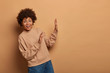 Joyful surprised woman raises palms towards brown background, steps aside from something, impressed by incredible thing, wears brown sweater and jeans. Blank space for your advertising content