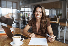 Business, Finance And Entrepreneur Concept. Happy Cheerful Attractive Spanish Woman Sitting Alone Cafe, Co-working Space With Opened Laptop, Working Remote, Drink Coffee And Read Paper, Studying