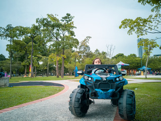 An Asian kid driving electric toy car in a tropical park. Outdoor toys. Children in battery power vehicle. Little girl riding toy truck in the garden. Family playing in the backyard.