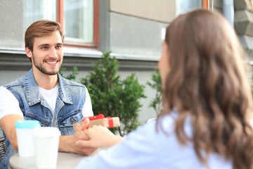 Wall Mural - Smiling young girl having a date with her boyfriend at the coffee shop, man holding present box.