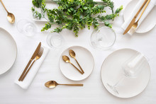 Tableware Background In White Gold Tone.White Plates,  Gold Cutlery,  Wine Glasses And Decorative Plant Centrepiece