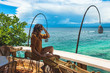 A young woman sits at the bar and enjoys the view of the endless blue sea and blue sky, Bali, Indonesia. Girl admires the view of the sea on the island of Bali