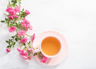 Cup of tea and branch of small pink  roses on  white rustic table. Top view.