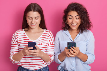 Horizontal shot of attractive Caucasian women standing isolated over pink background with phones in their hands, looking ar device's screens with happy axpressions, ladies wearing casual clothing.