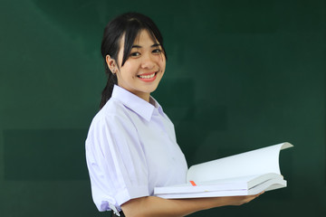 Smiling happy young portrait pretty Asian student girl holding book at green board in high school or college uniform, smile at front classroom for learning. Education learning in college