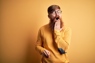 Canvas Print - Handsome Irish redhead man with beard wearing glasses over yellow isolated background bored yawning tired covering mouth with hand. Restless and sleepiness.