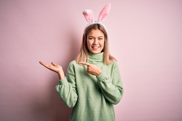 Poster - Young beautiful woman wearing easter rabbit ears standing over isolated pink background amazed and smiling to the camera while presenting with hand and pointing with finger.