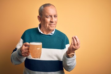 Sticker - Senior handsome man drinking jar of beer standing over isolated yellow background doing money gesture with hands, asking for salary payment, millionaire business