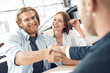 © Viktoriia - Car Salon. Young couple sitting at table man shaking hands with agent buying car while woman holding keys happy