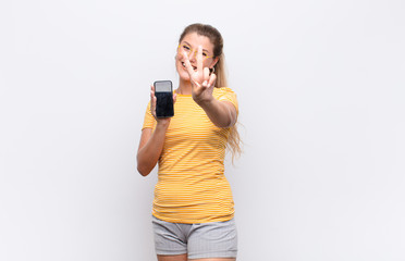 young pretty latin woman smiling and looking friendly, showing number two or second with hand forward, counting down with a smartphone against white wall